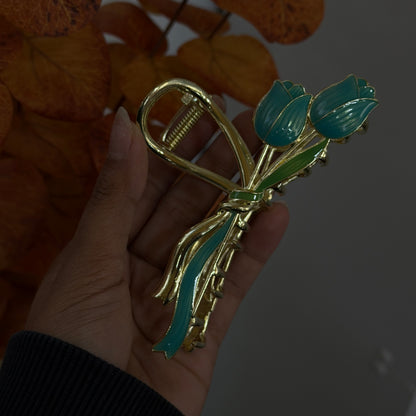 Gold hair claw designed as a bow with teal tulip flowers and green leaves, finished with glossy enamel details and shown held in hand.