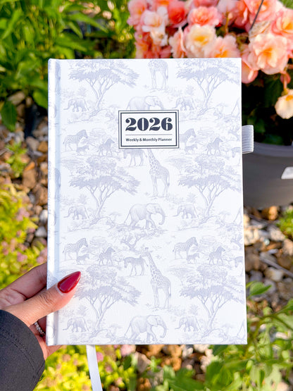 White hardcover 2026 weekly & monthly planner with safari animal pattern, held in hand with red nails, floral background