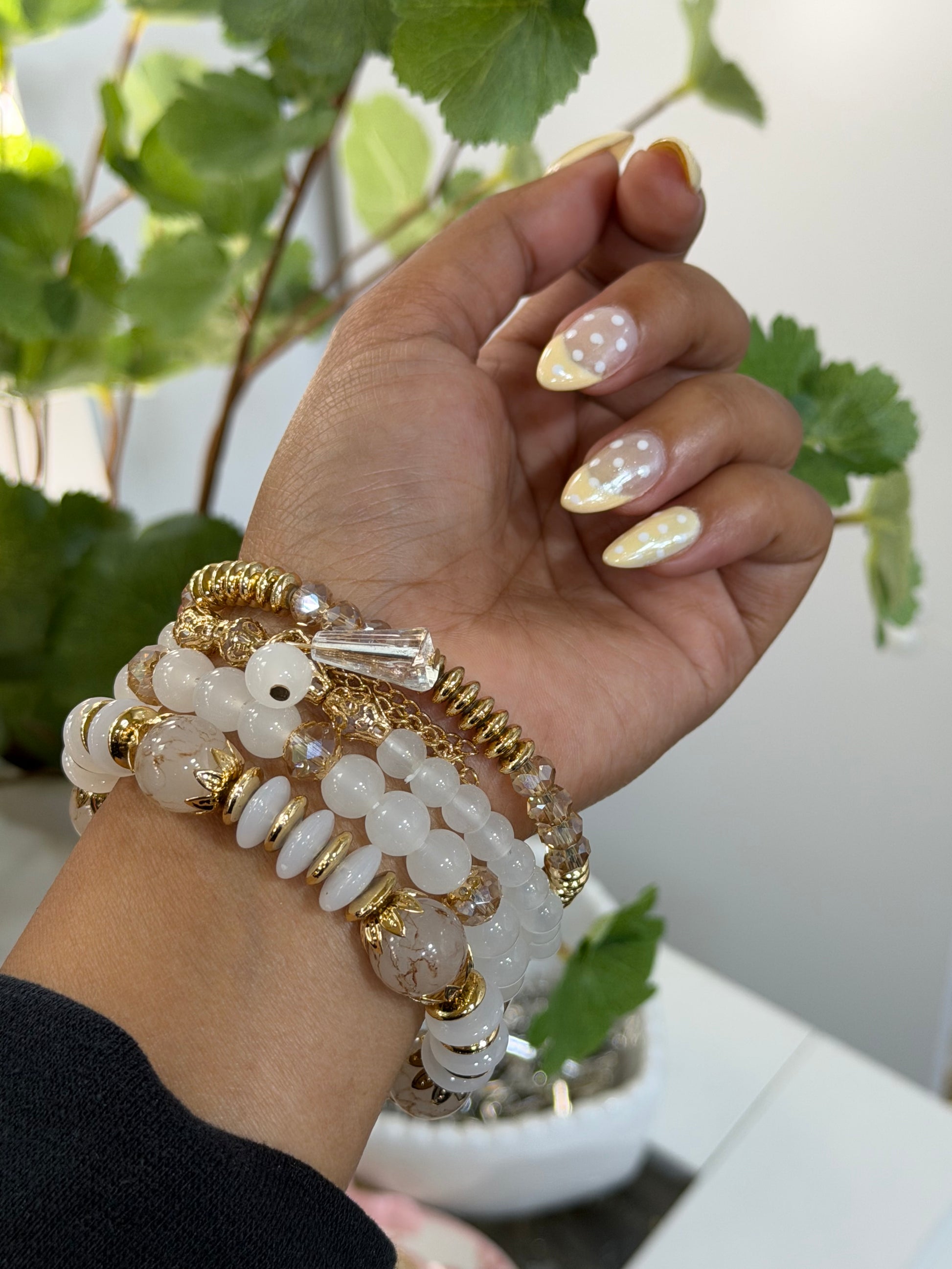 Layered beaded charm bracelets with white and gold beads and clear crystal accents, worn on a hand with yellow polka dot nails.