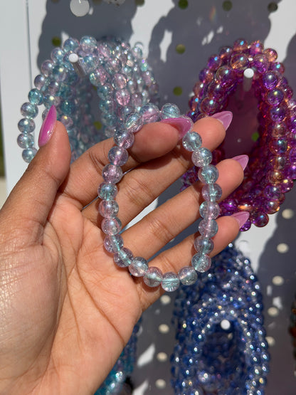 Blue beaded bracelets, including one iridescent blue and pink, held by a hand against a display of other colorful beaded bracelets.