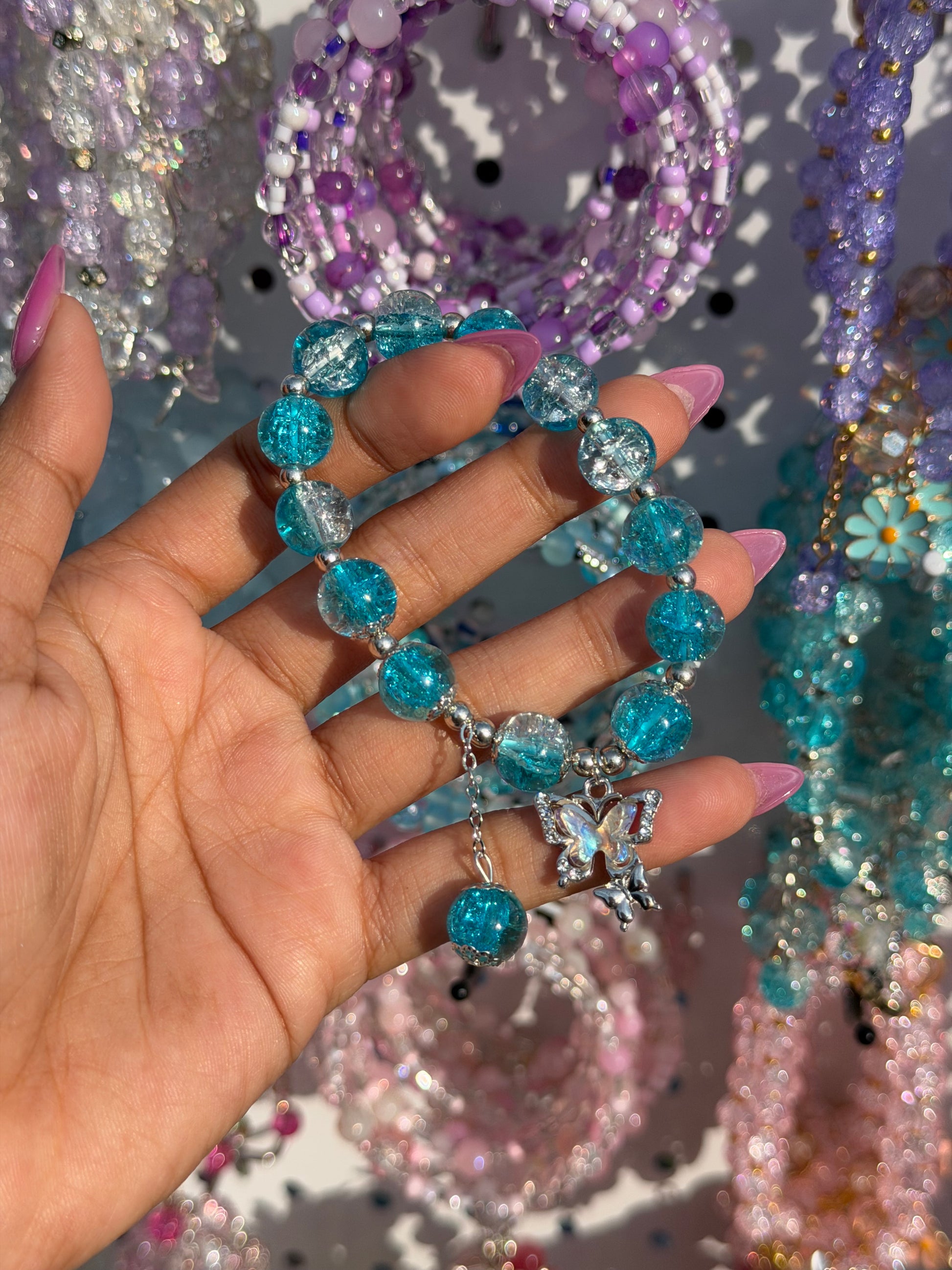 Blue and clear cracked glass beaded bracelet with a silver butterfly charm, held by a hand with pink nails against a blurred background of colorful beads.