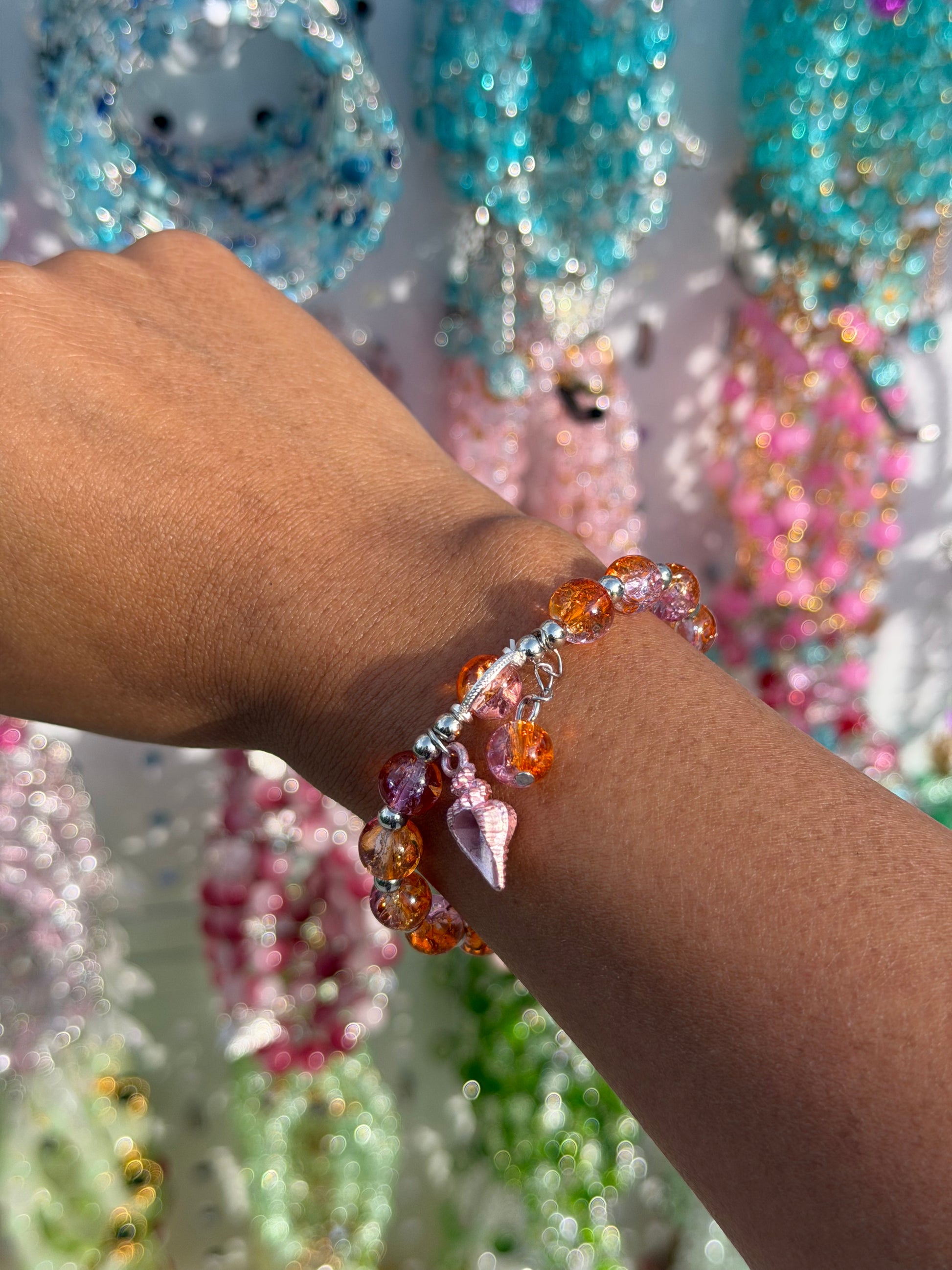 Beaded bracelet with orange and pink crackled beads and a pink shell charm, worn on a wrist.