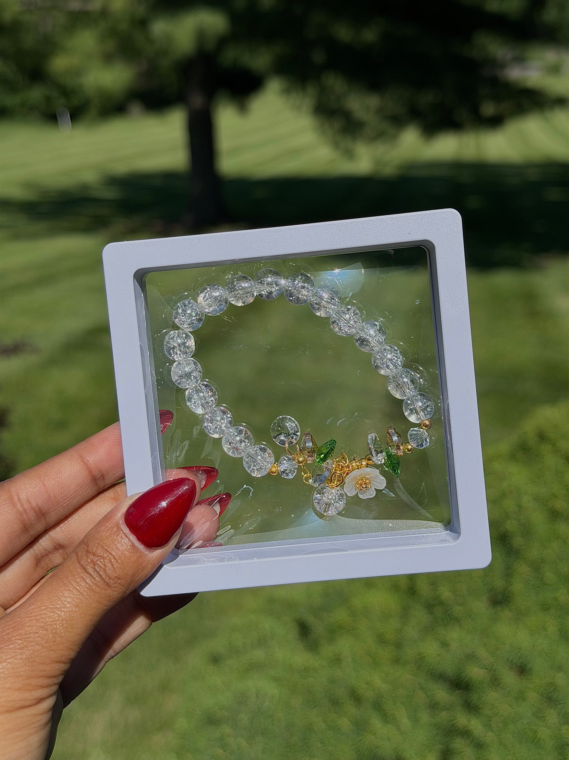 Clear beaded bracelet with floral charm and gold accents in white display case, held in hand with red nail polish, outdoor grass background