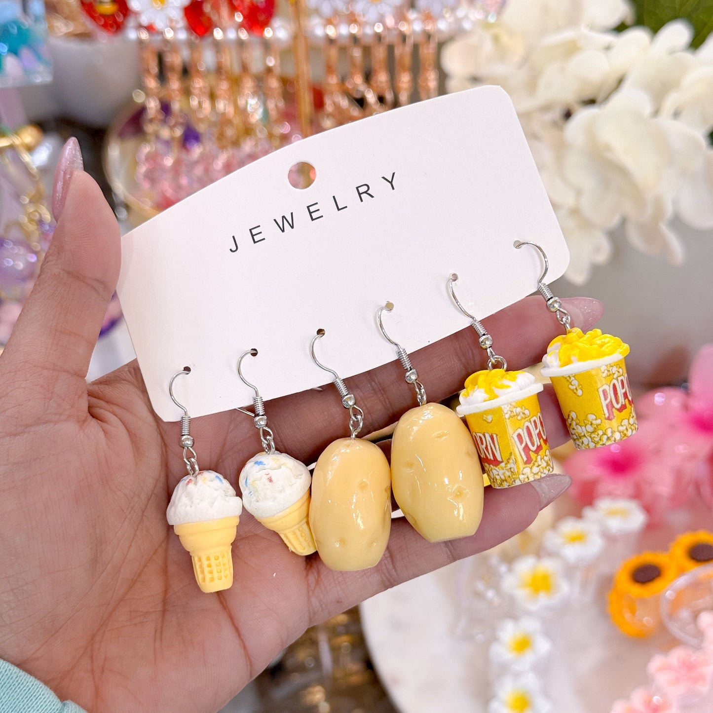 Hand holding a jewelry card displaying five novelty dangling earrings: two yellow ice cream cone earrings, two yellow potato-shaped earrings, and one yellow popcorn bucket earring with popped popcorn details. 