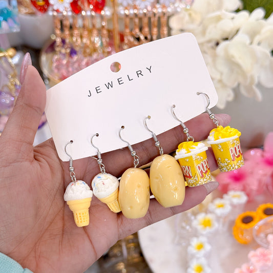 Hand holding a jewelry card displaying five novelty dangling earrings: two yellow ice cream cone earrings, two yellow potato-shaped earrings, and one yellow popcorn bucket earring with popped popcorn details. 