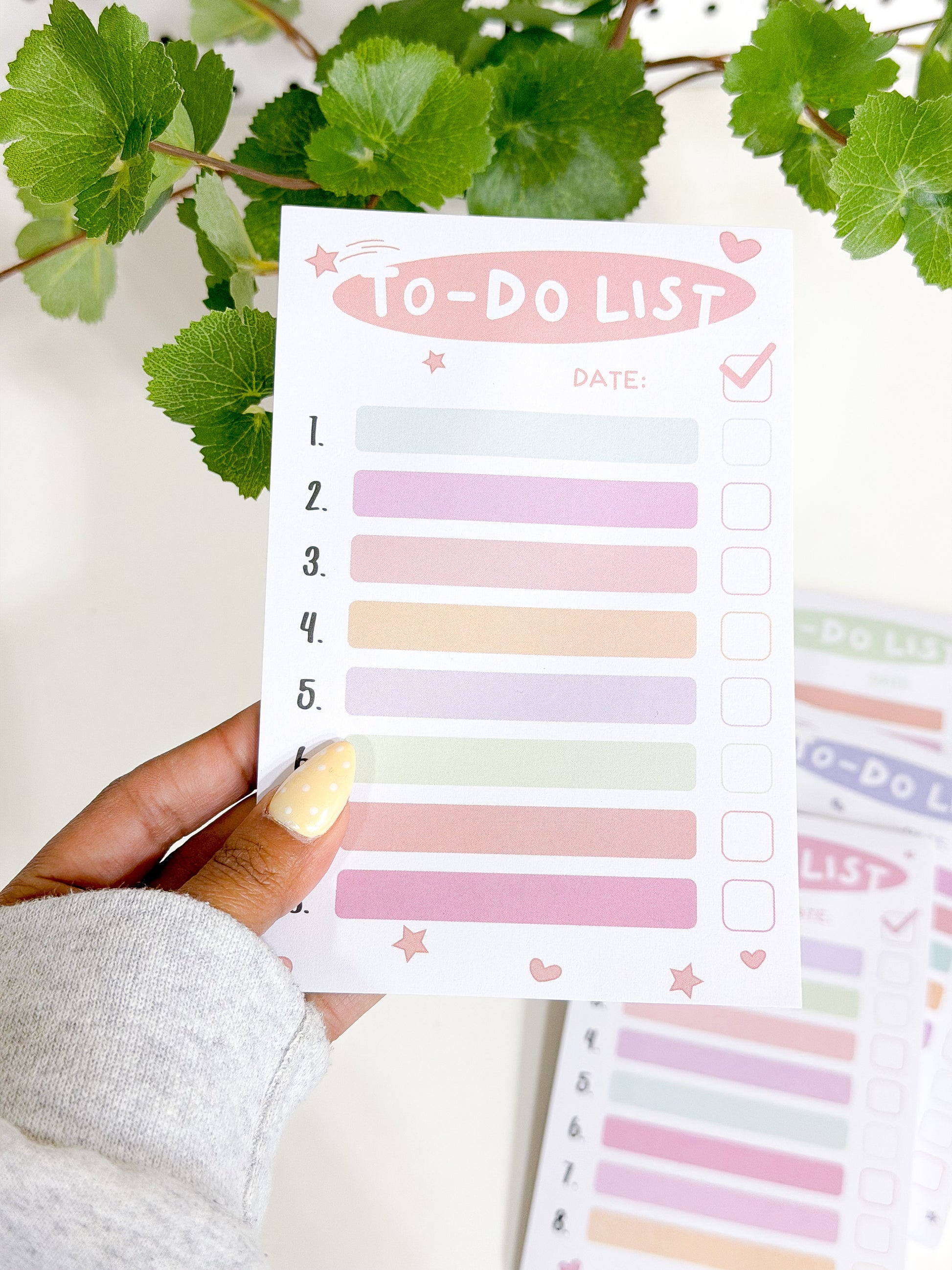 White Tasks Notepad with numbered pastel lines and checkbox squares, decorated with pink stars and hearts, held in a hand.