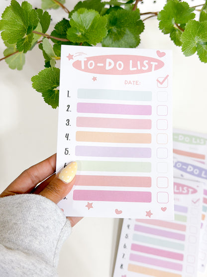 White Tasks Notepad with numbered pastel lines and checkbox squares, decorated with pink stars and hearts, held in a hand.