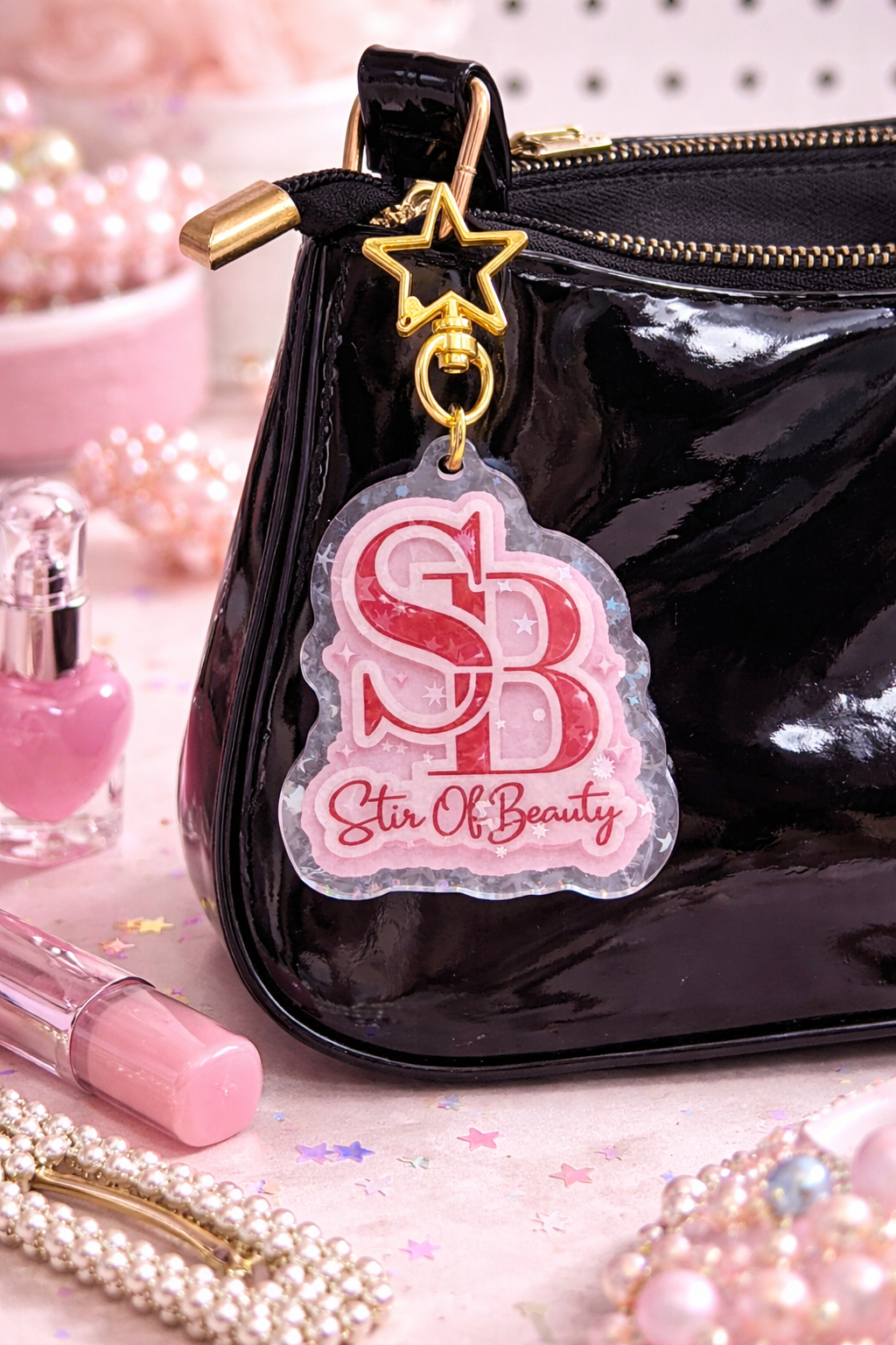Pink and white keychain charm by Stir Of Beauty with SB logo and text, attached to a gold star clasp on a black bag.