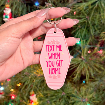 Pink motel-style keychain with "Text me when you get home" text, in front of a blurred Christmas tree.
