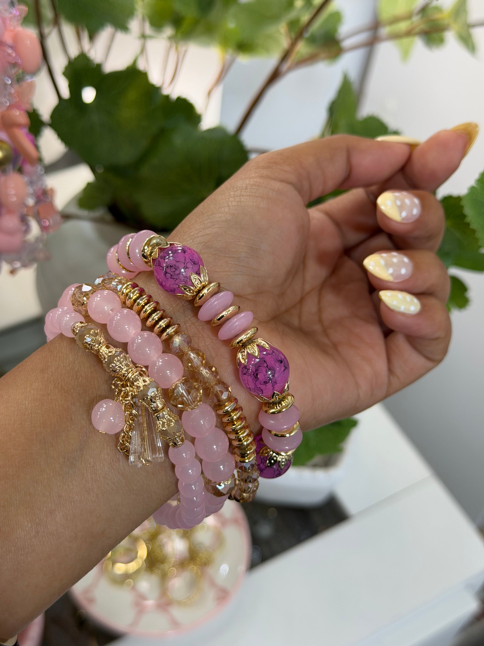 Layered pink and gold beaded charm bracelets with purple marbled beads and a clear tassel, worn on a wrist with yellow polka dot nails.