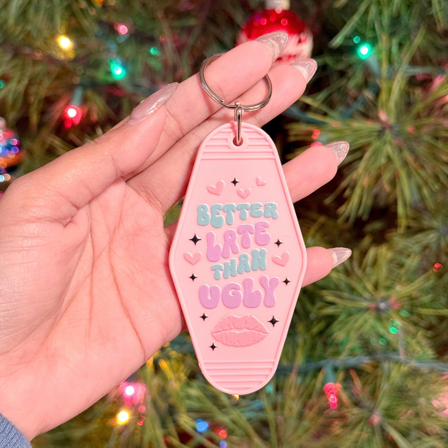 Pink motel keychain with "Better Late Than Ugly" by an unspecified brand, held in a hand against a blurry Christmas tree.
