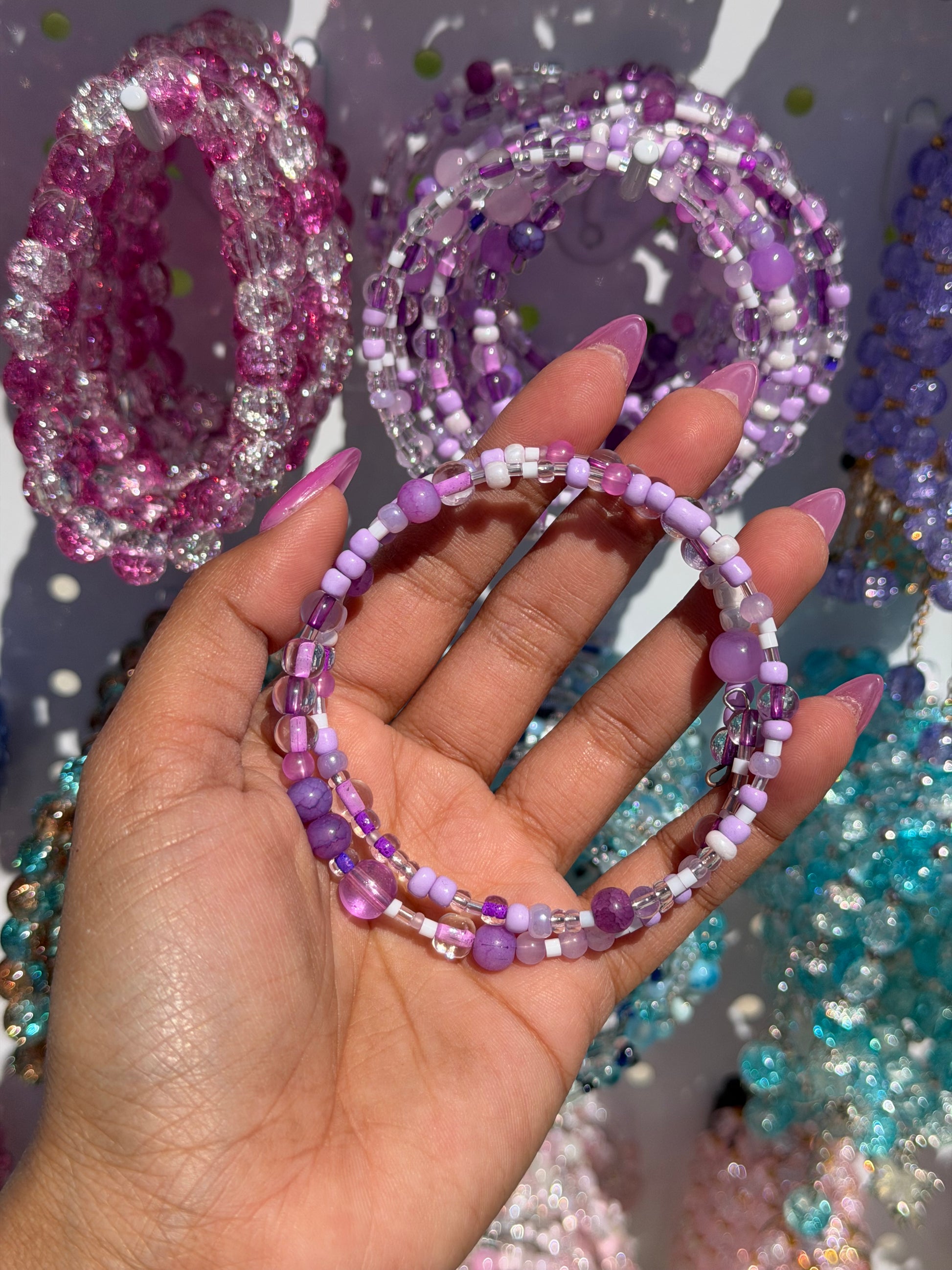 Layered beaded bracelets with purple, white, and clear beads, held by a hand with long pink nails.