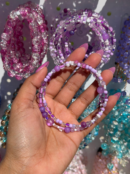 Layered beaded bracelets with purple, white, and clear beads, held by a hand with long pink nails.