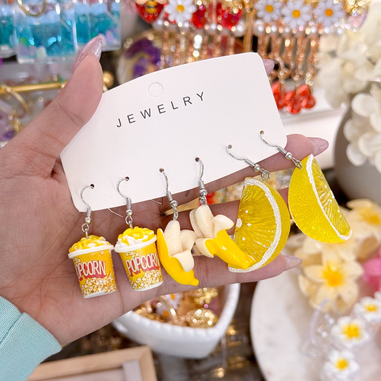 Hand holding a jewelry card with novelty dangling earrings shaped like popcorn buckets, peeled bananas, and yellow lemon slices on silver hooks.