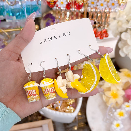 Hand holding a jewelry card with novelty dangling earrings shaped like popcorn buckets, peeled bananas, and yellow lemon slices on silver hooks.