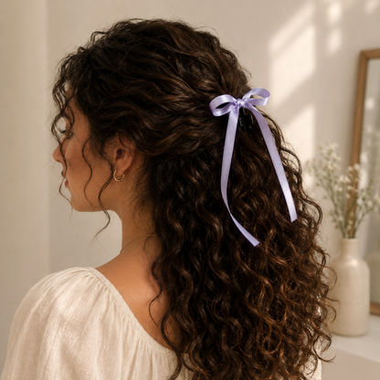 Lavender bow mini hair clip holding back dark curly hair, seen from the side on a woman wearing a white top.