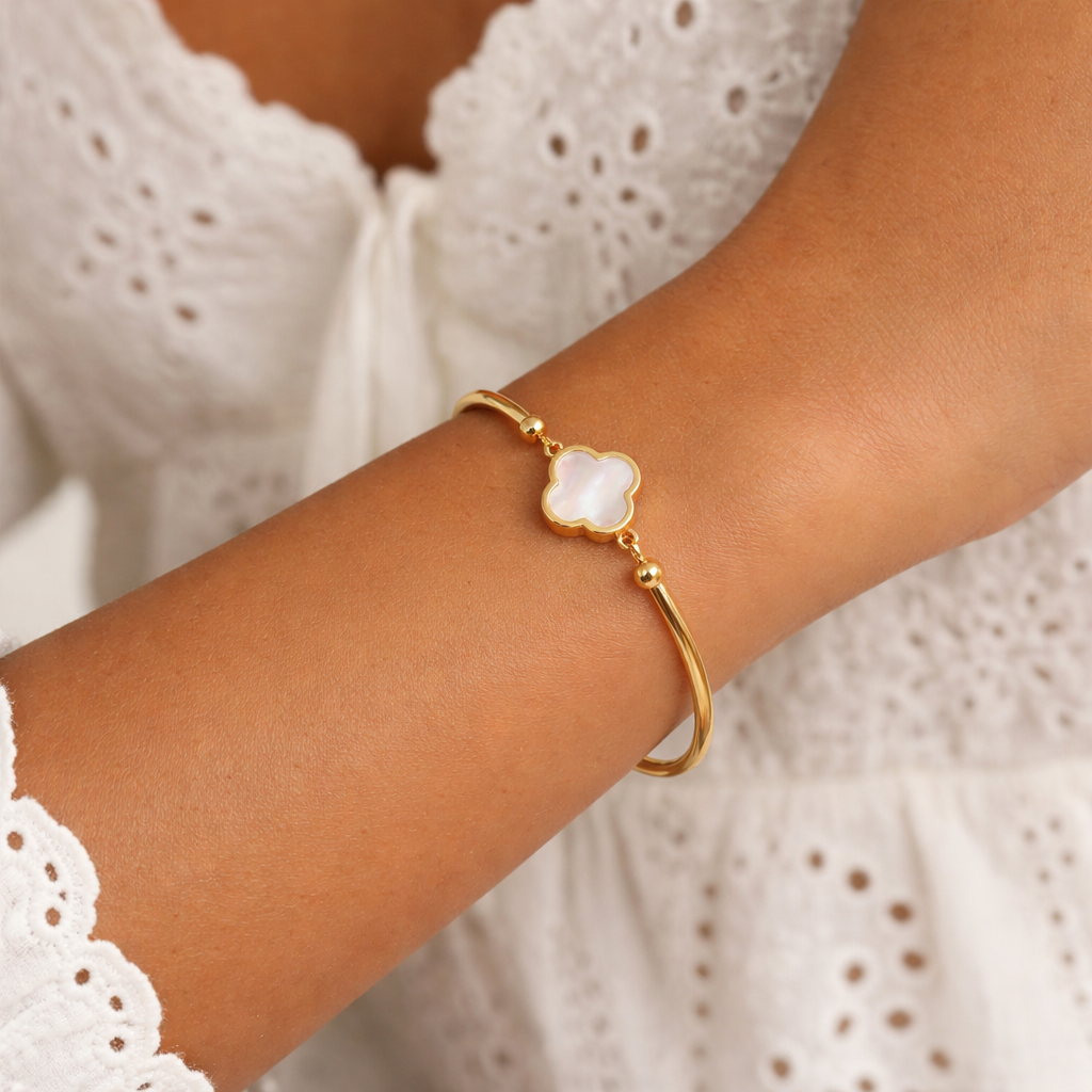Gold-colored bangle bracelet featuring a white clover charm, worn on a woman's arm with a white lace top.