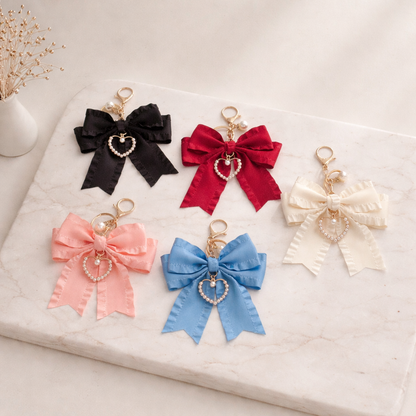 Five colorful satin Pearl Bow Bag Charms with pearl heart pendants and gold-tone hardware, displayed on a marble surface.