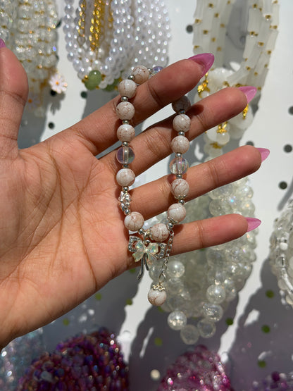 White beaded bracelet held by a hand with pink nails, featuring cracked-look beads, clear beads, and a silver bow charm.