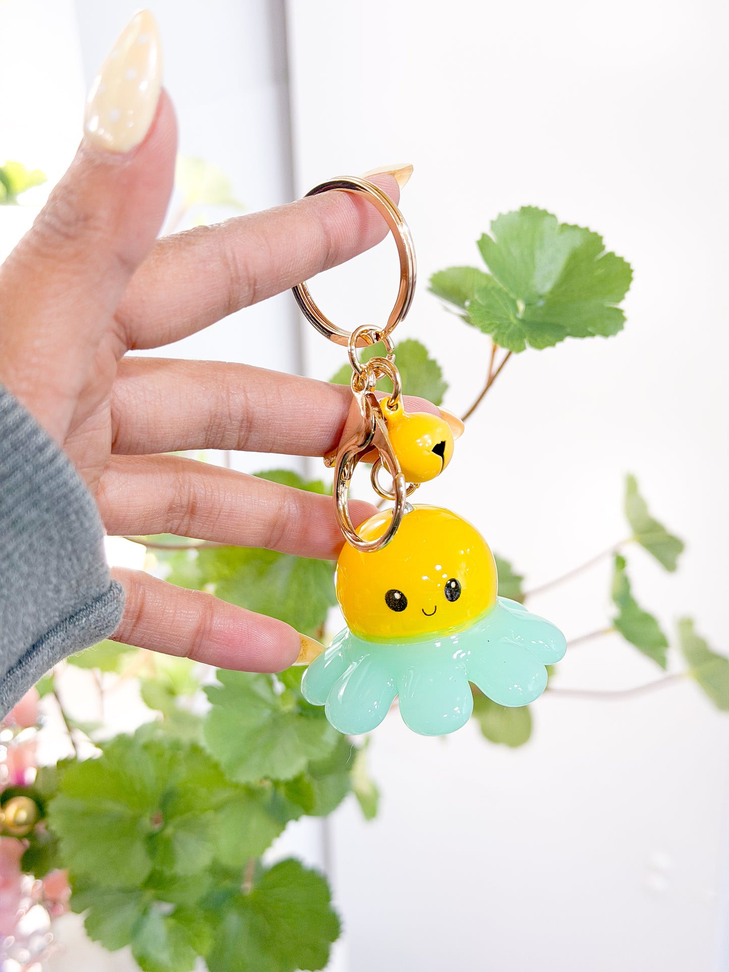 Yellow and mint green glossy octopus charm with a smiling face, attached to a gold keyring and small yellow bell, held by a hand.