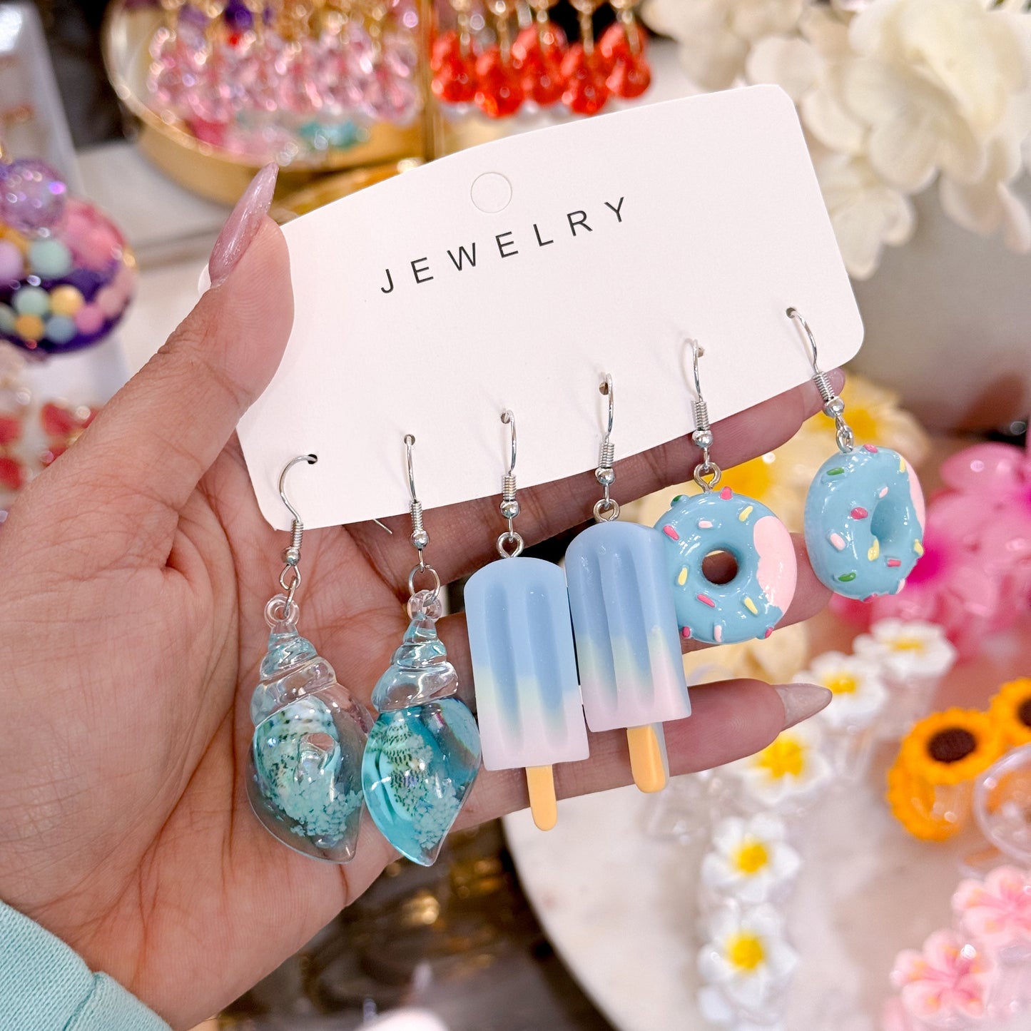 Hand holding a jewelry card with novelty dangling earrings including blue seashell charms, light blue popsicle earrings, and blue donut charms with sprinkles, displayed on silver hooks.