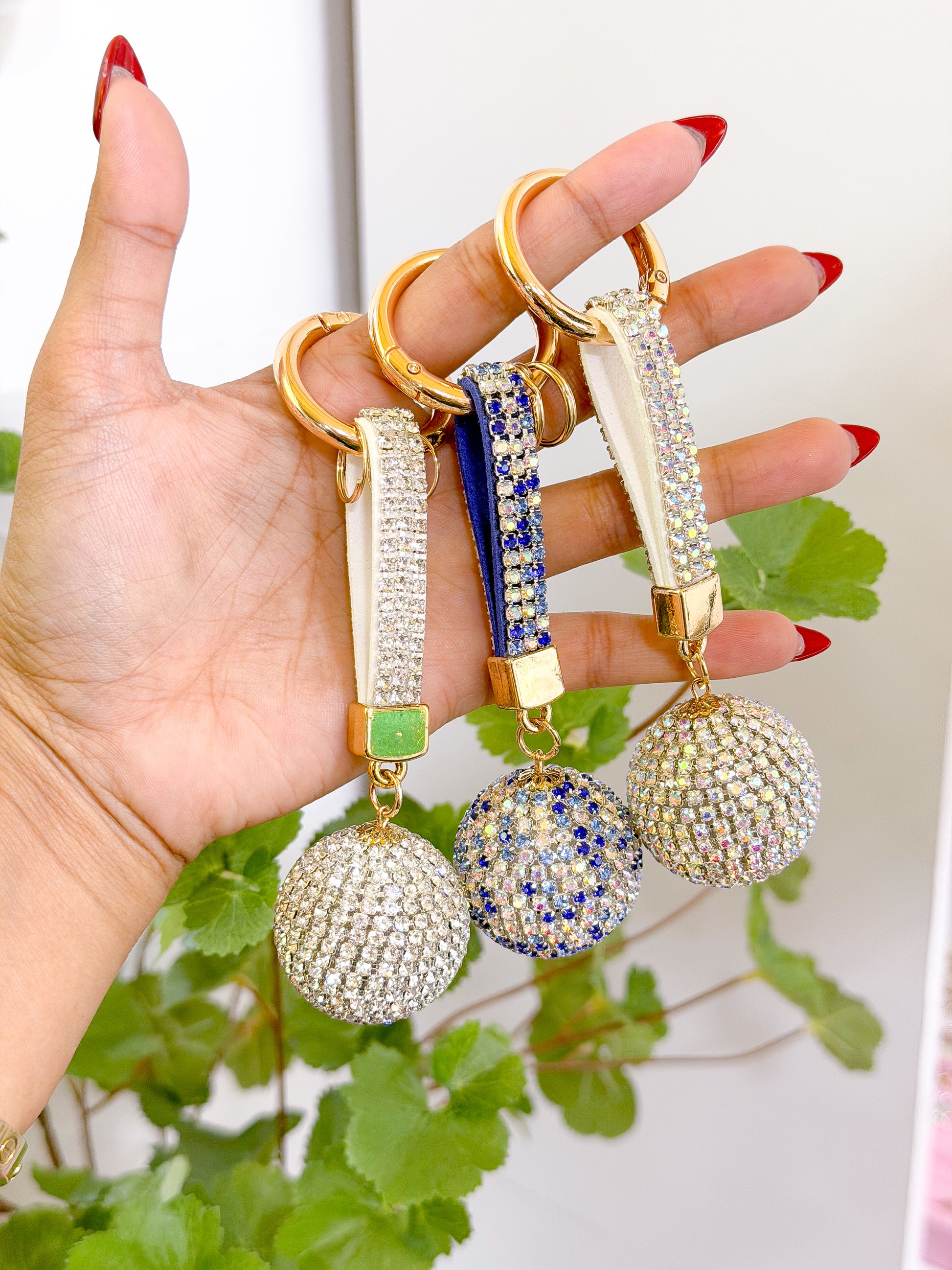 Shiny rhinestone disco ball bag charms in silver, blue, and clear, held in hand with gold rings and velvet straps, green leaf background