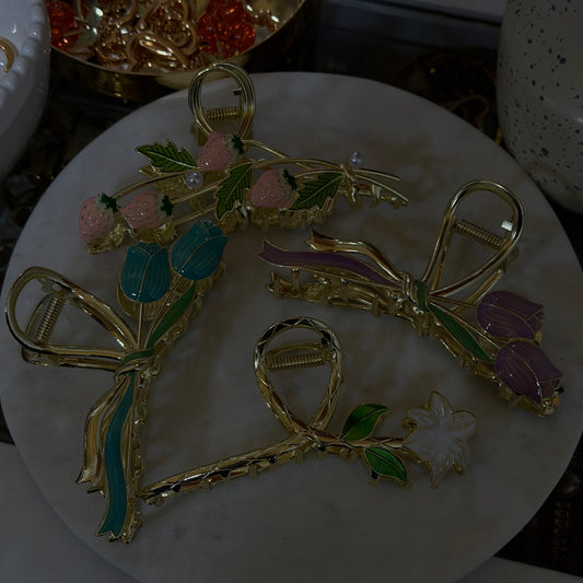 Four floral gold hair claws featuring strawberry, teal tulip, purple tulip, and white flower designs arranged together on a neutral surface.