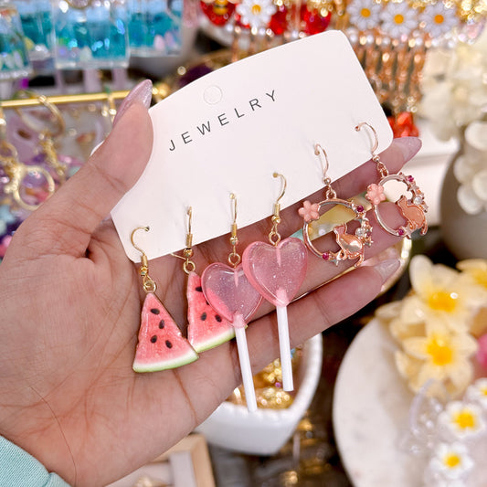 Hand holding a jewelry card with novelty dangling earrings including pink watermelon slice charms, translucent heart-shaped lollipop earrings, and pink floral hoop earrings on gold hooks.