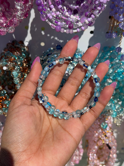 Layered blue, light blue, and clear beaded bracelet with silver and black accents, held by a hand with pink nails.