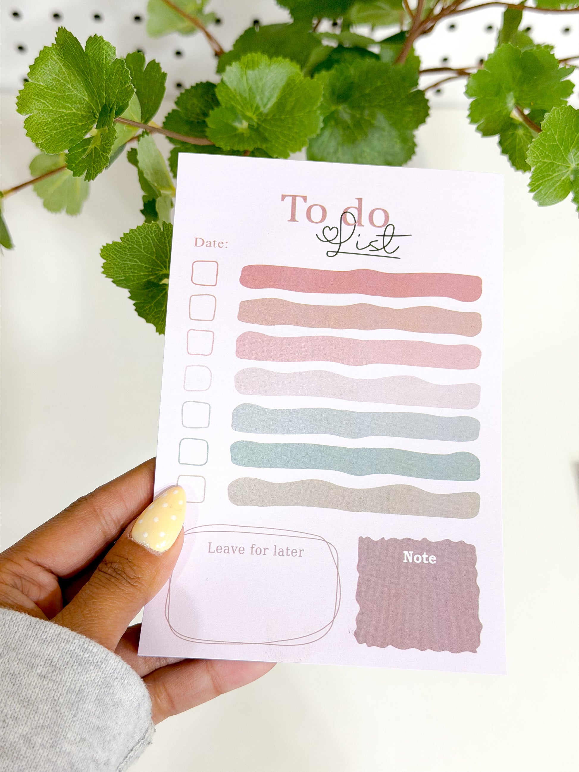 Pink pastel colorful striped To Do List Notepad held by a hand with a yellow polka dot nail, against green leaves.