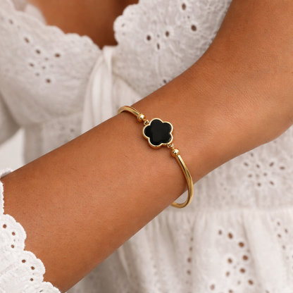 Gold bangle bracelet with a black clover centerpiece, worn on a woman's arm with a white embroidered top.