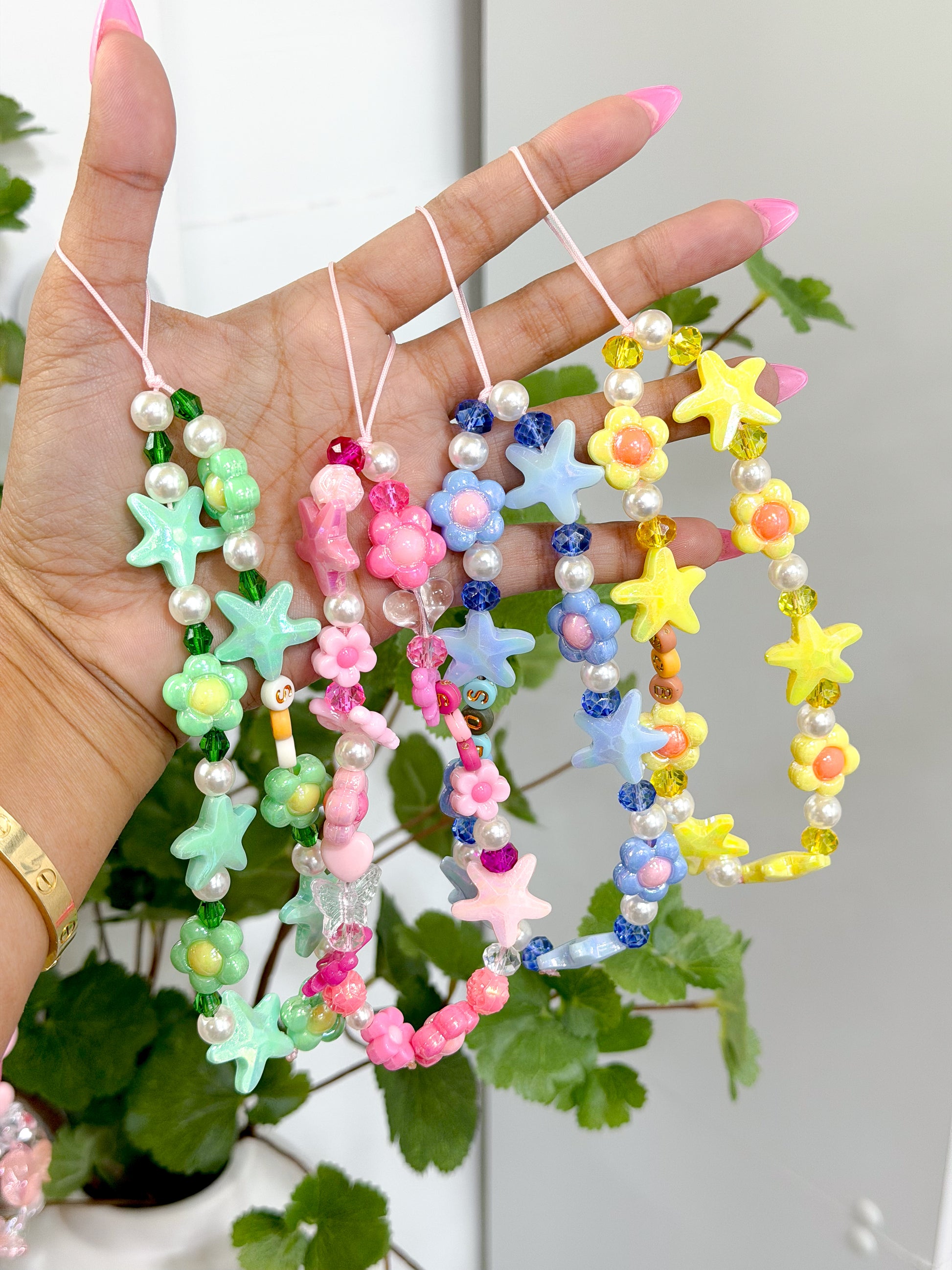 Vibrant multi-colored beaded phone charms by SOB with star, flower, and letter beads, held by a hand with pink nails and a gold bracelet.