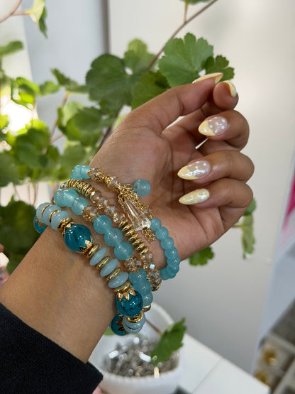 Layered blue and gold beaded charm bracelets with a clear faceted charm, worn on a hand with yellow polka dot nails.