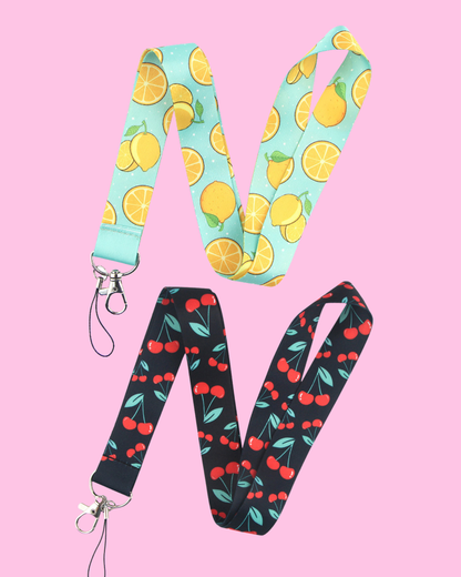 Two phone lanyards, one light blue with yellow citrus fruit and the other black with red cherries, both having silver clasps.