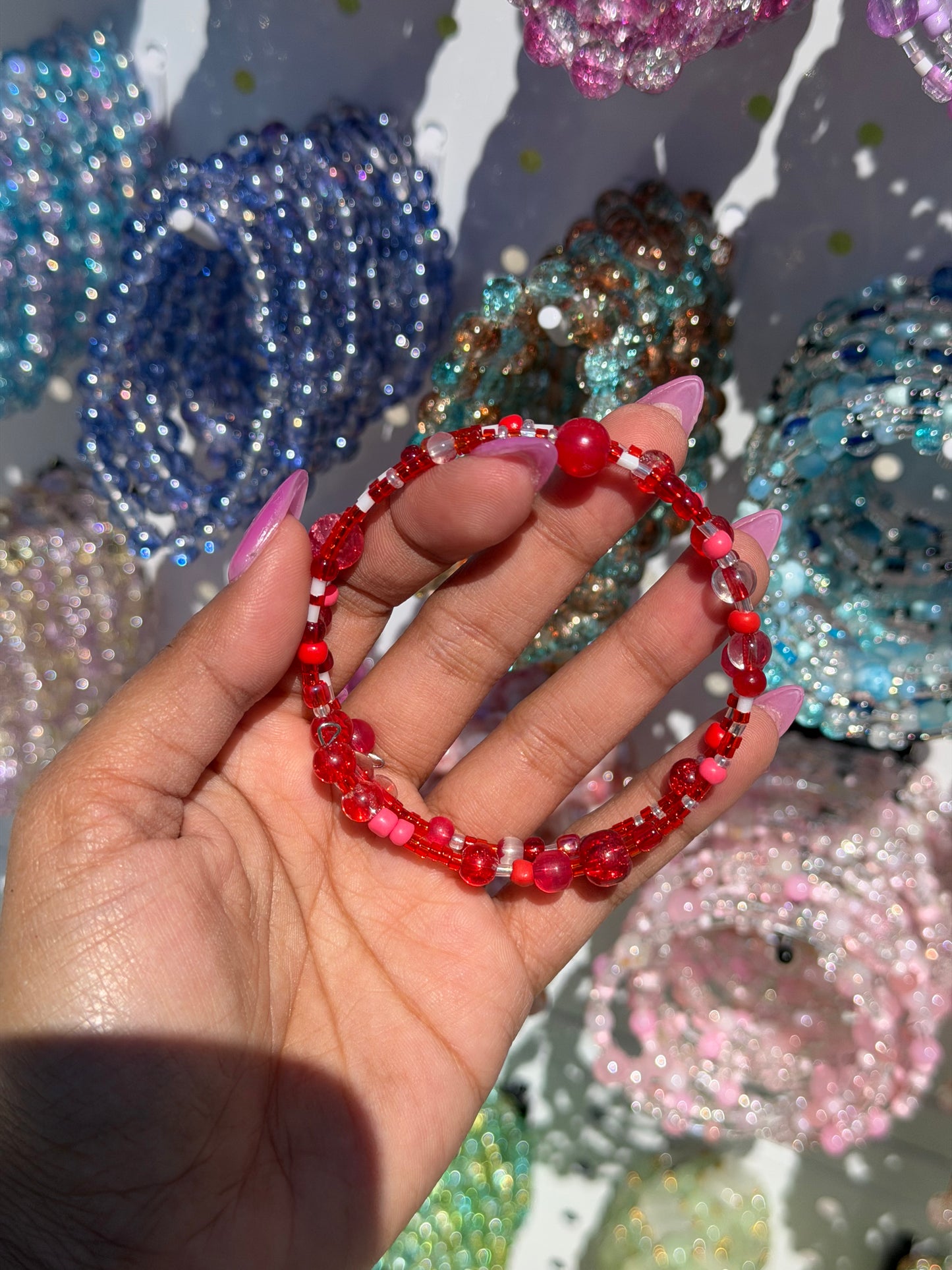 Layered red beaded bracelets held by a hand with pink nails, with other colorful beaded bracelets blurred in the background.