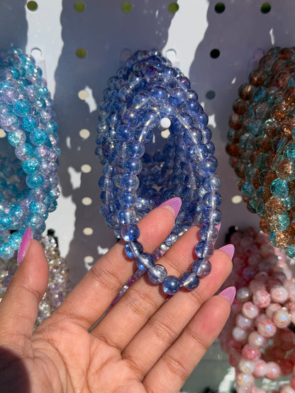 Blue beaded bracelets held by a hand, showing sparkling crackle beads alongside other colorful styles.