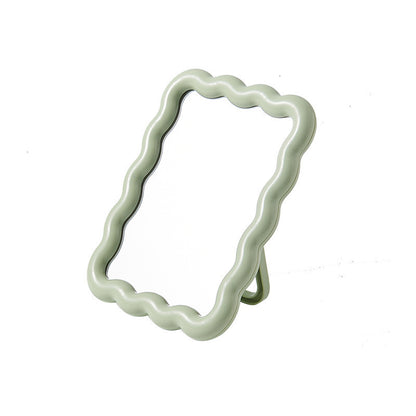 Light sage green ripple tabletop mirror with a wavy-edge frame and kickstand, angled view.