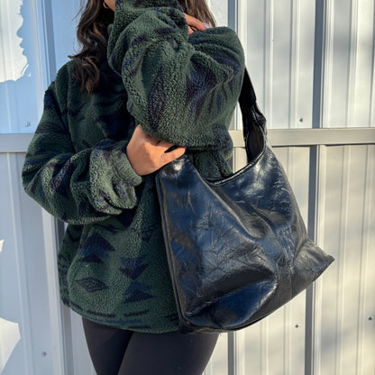 Black leather tote bag with shoulder strap, styled with a green patterned fleece jacket and black leggings.
