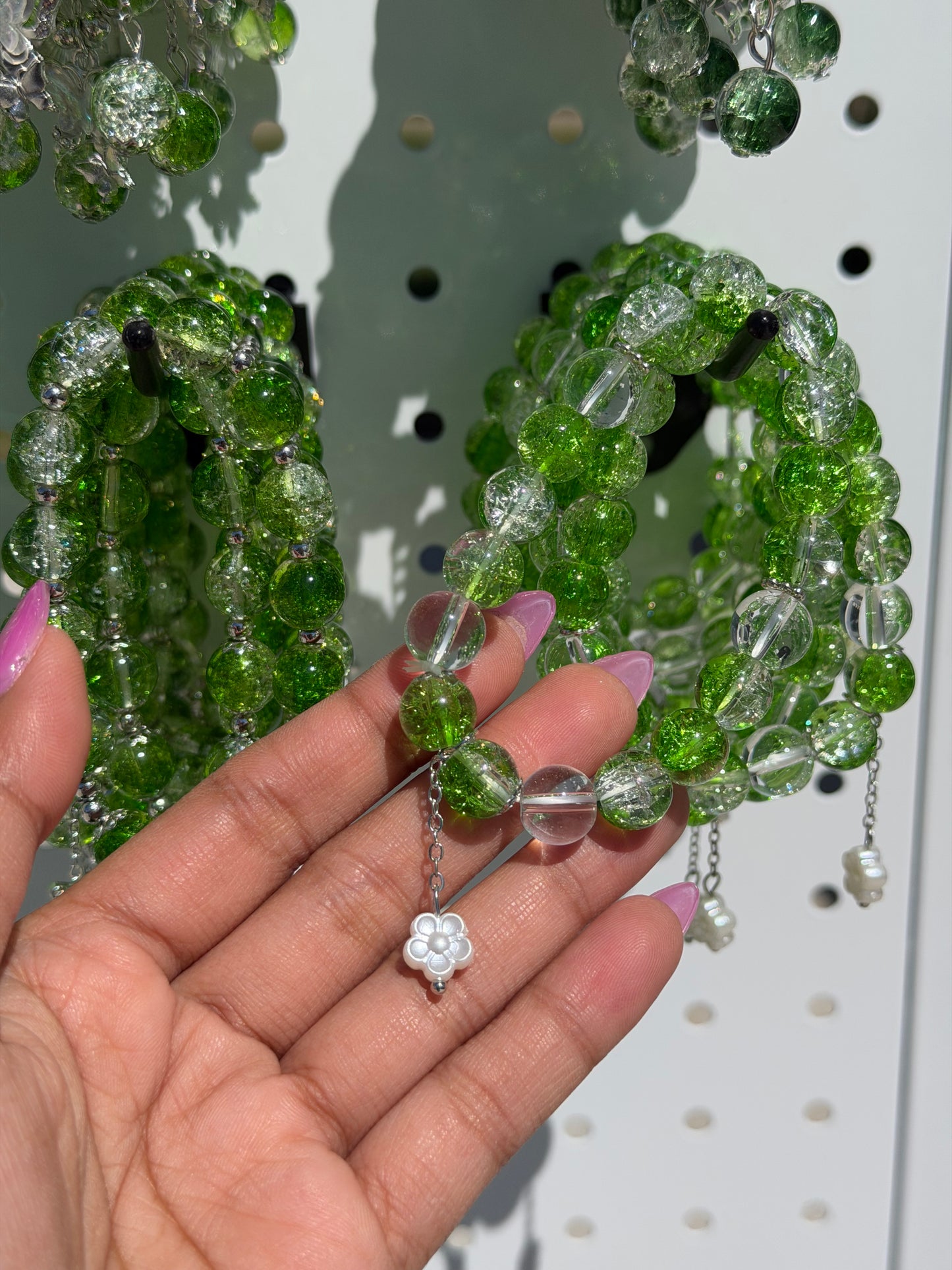 Green and clear beaded bracelets held by a hand, featuring cracked and smooth beads, silver spacers, and a white flower charm.