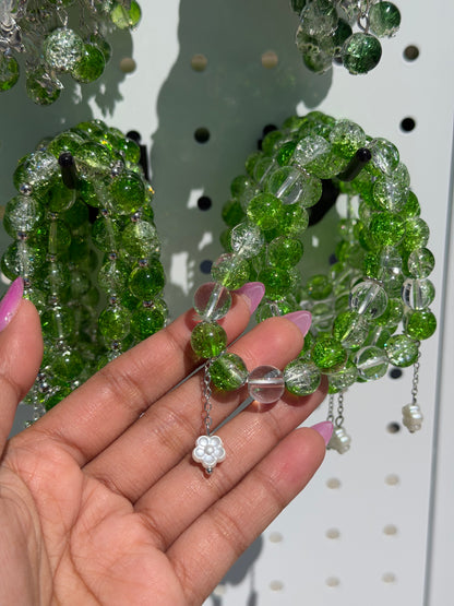 Green and clear beaded bracelets held by a hand, featuring cracked and smooth beads, silver spacers, and a white flower charm.