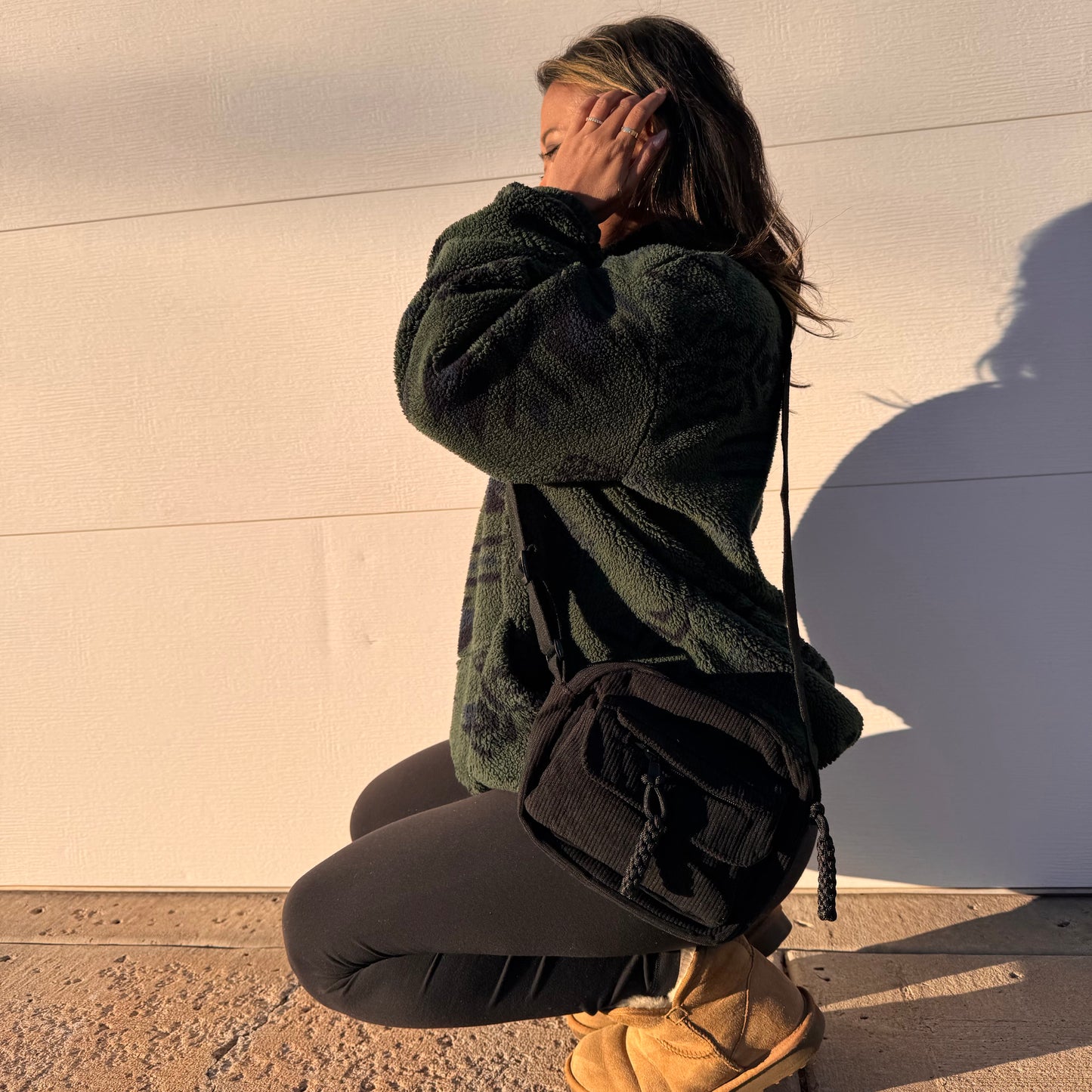 Black corduroy crossbody bag from a vendor, with a front pocket, styled with a green jacket, leggings, and boots.