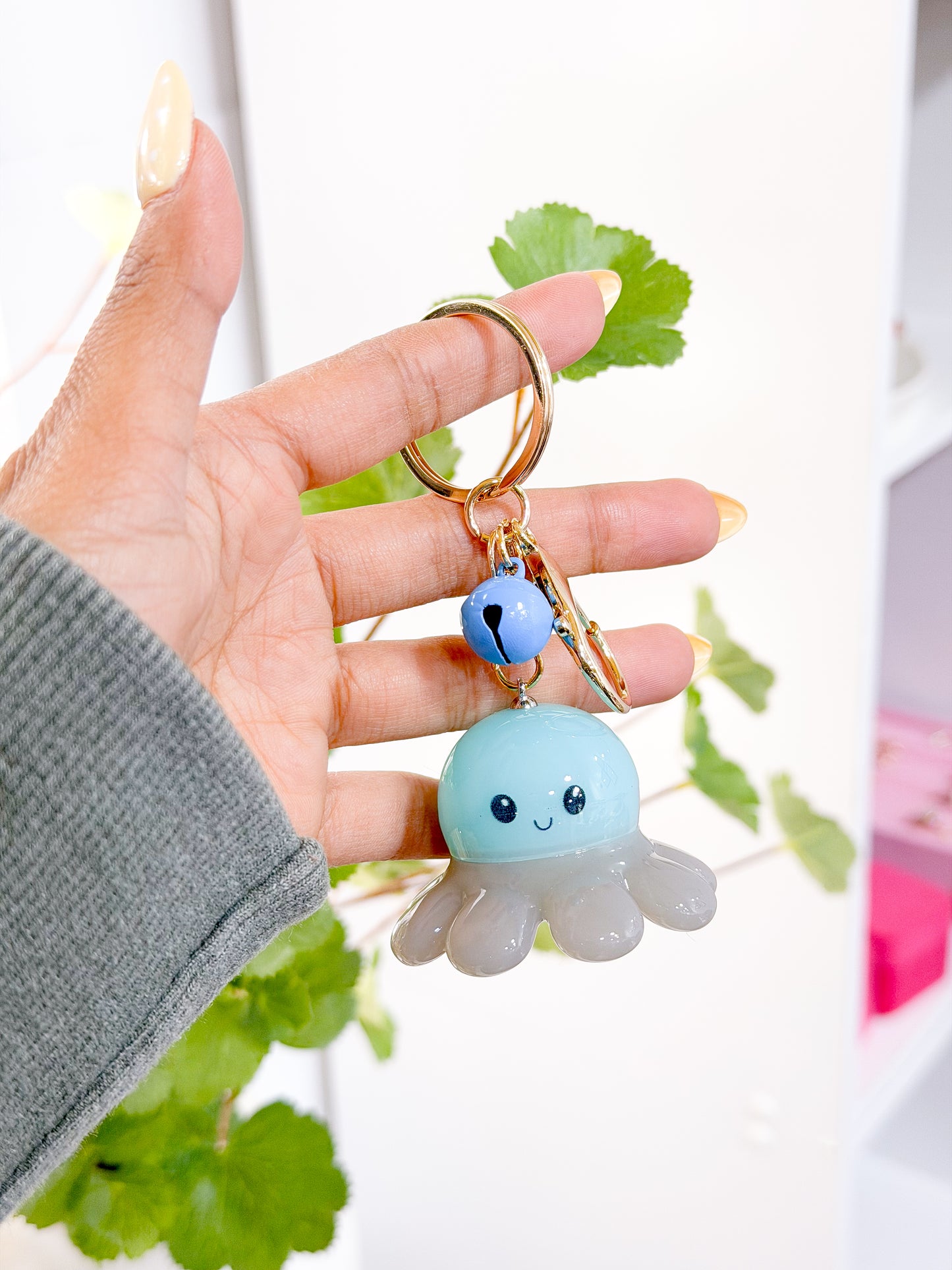 Light blue and translucent gray Glow Baby Octopus Charm with a smiling face, attached to a gold keychain with a blue bell, held by a hand.