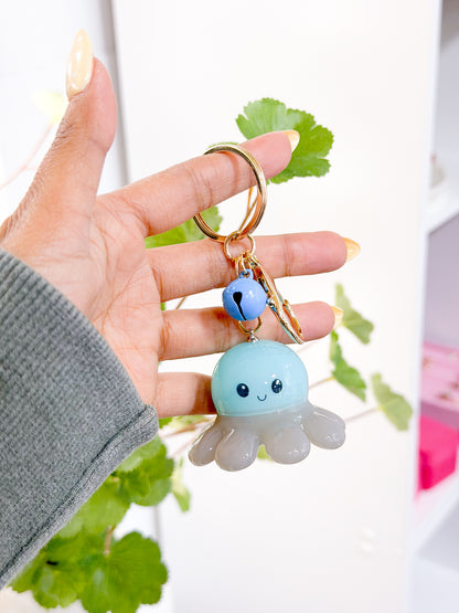 Light blue and translucent gray Glow Baby Octopus Charm with a smiling face, attached to a gold keychain with a blue bell, held by a hand.