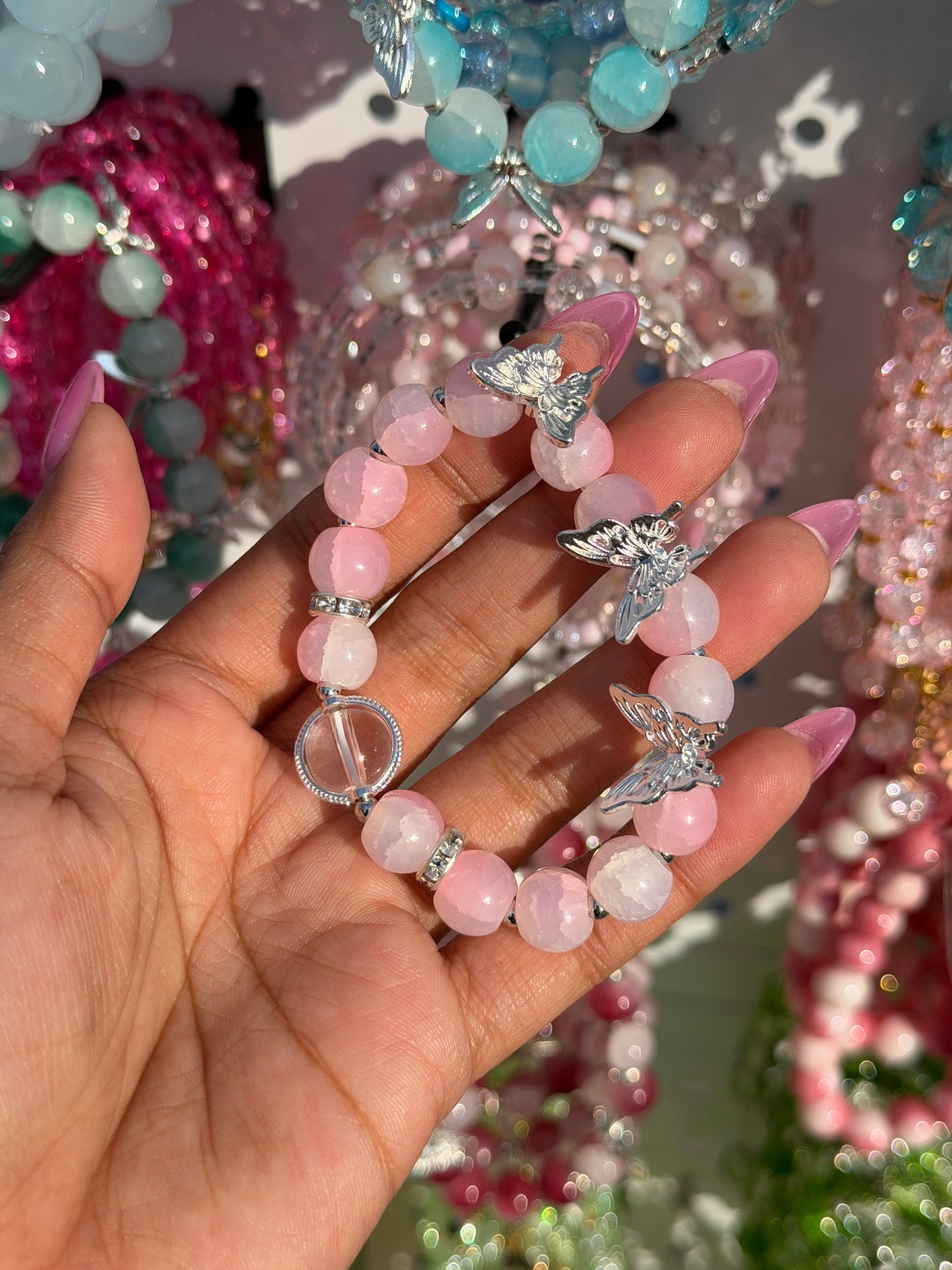 Pink beaded bracelet with silver butterfly charms, held by a hand with pink nails against a blurred backdrop of other jewelry.