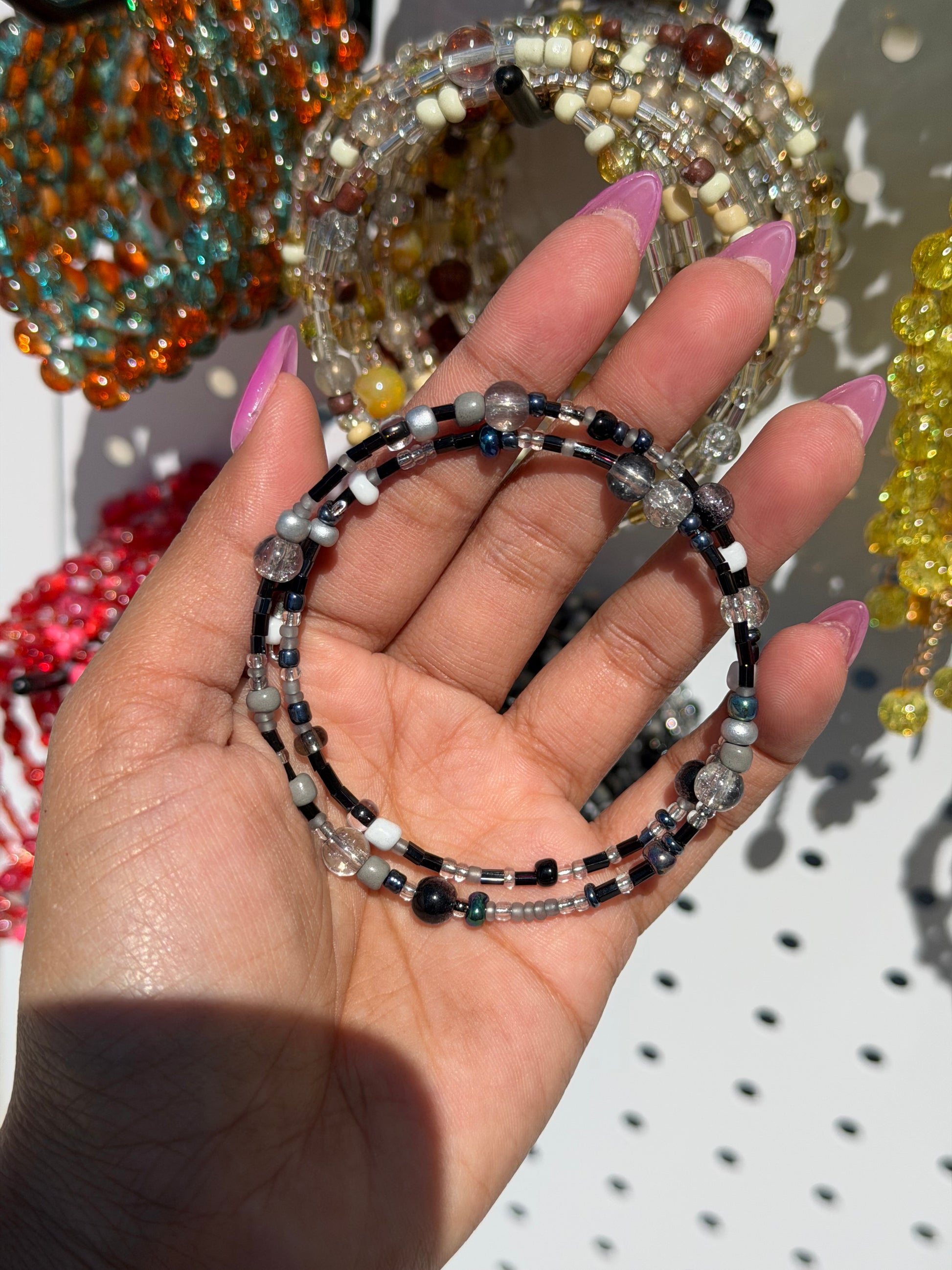 Layered beaded bracelets in black, white, grey, and clear, worn on a hand with pink nails, against blurry colorful jewelry.