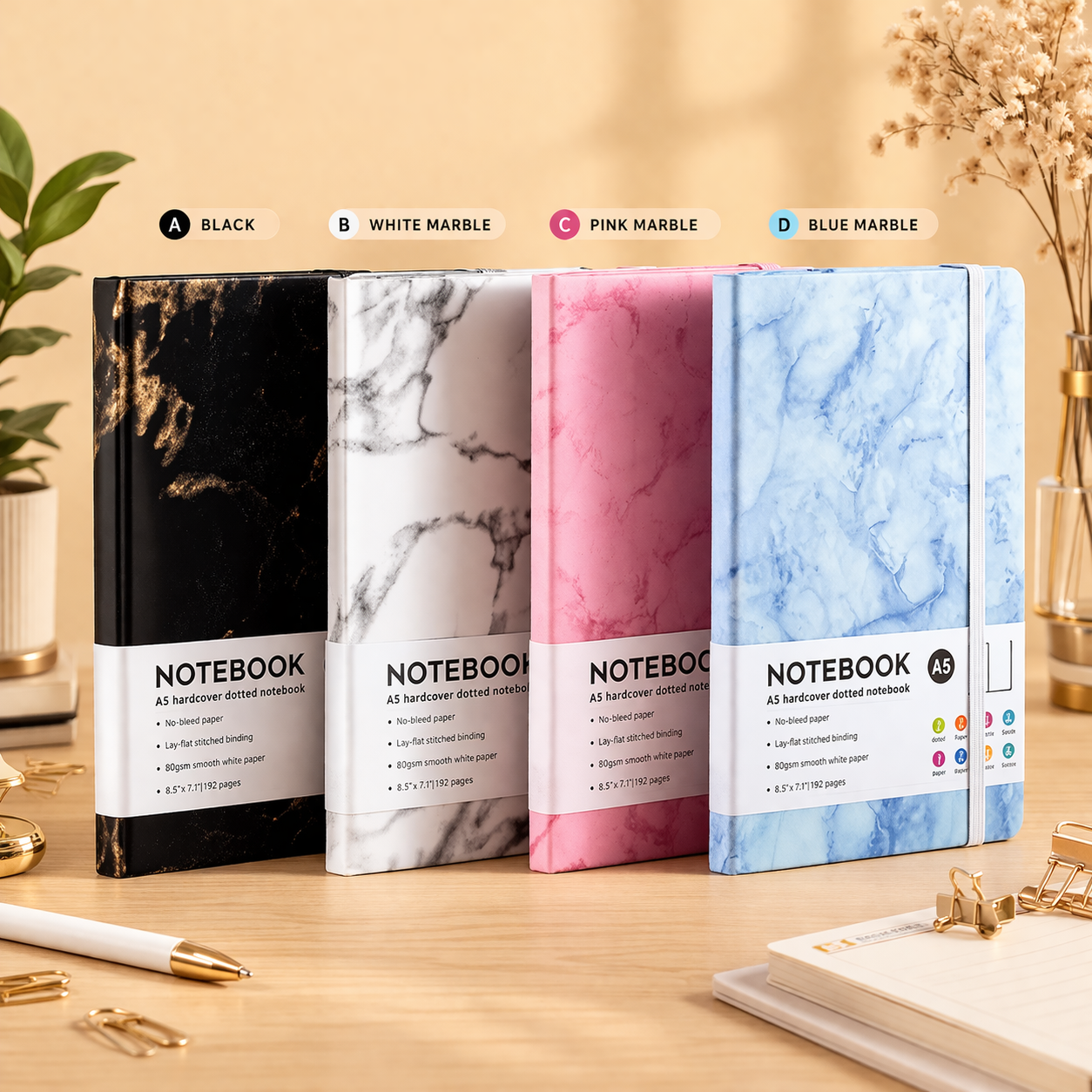 Four A5 hardcover dotted notebooks in black, white, pink, and blue marble patterns, lined up on a wooden desk with a pen and paper clips.