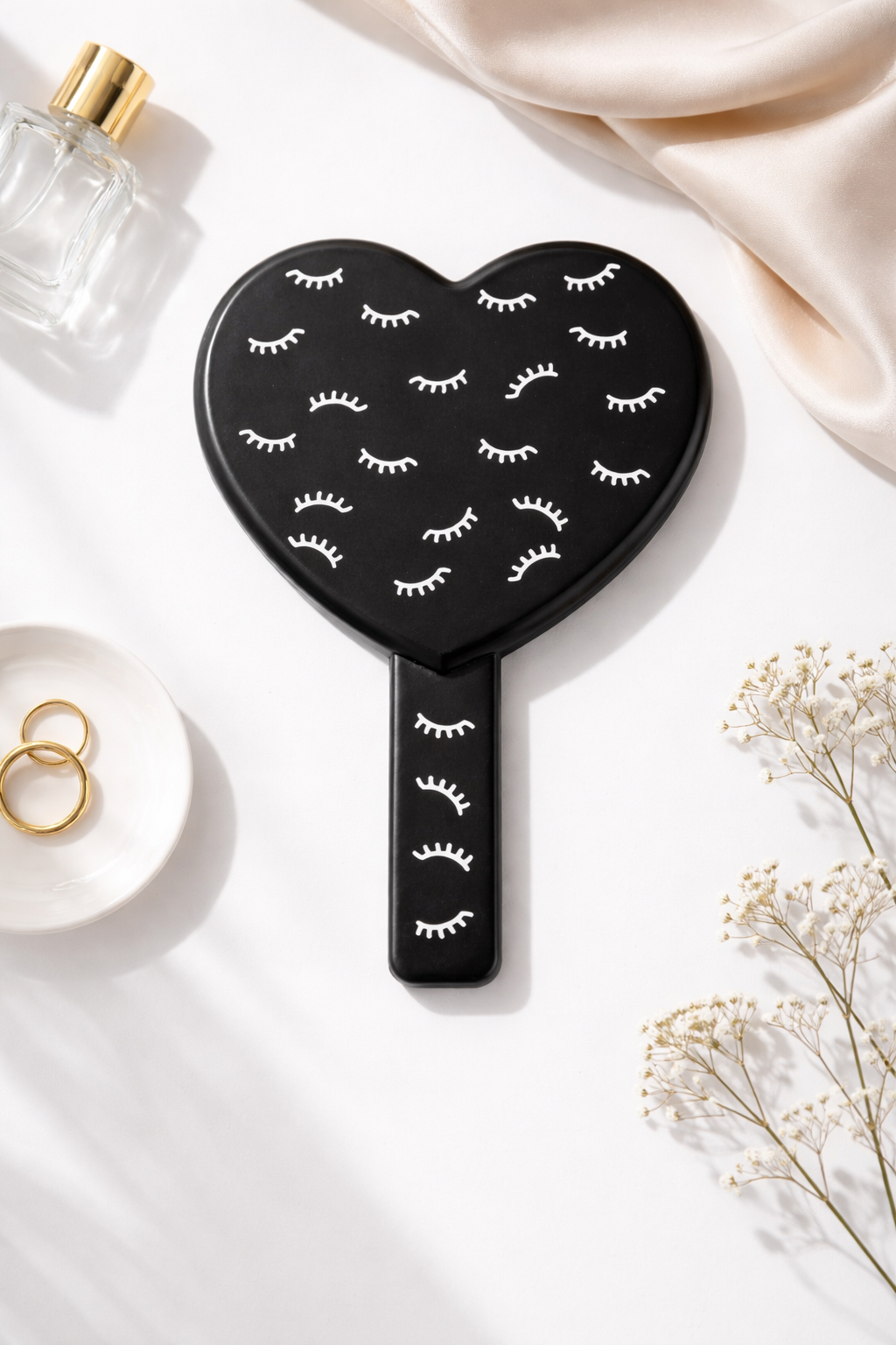 Black heart-shaped mirror with white eyelash patterns, styled with a perfume bottle, gold rings, and white baby's breath.