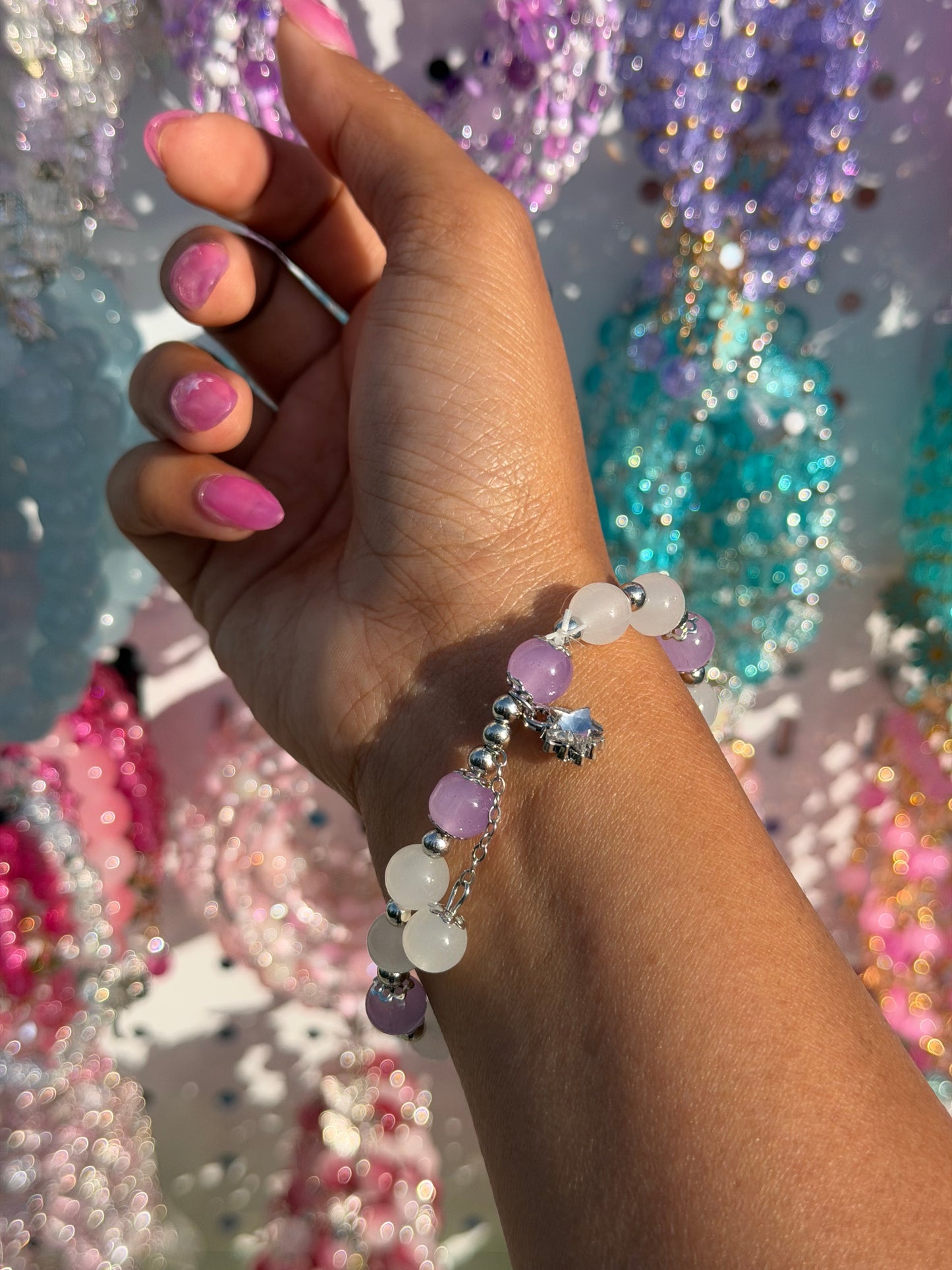 Lilac and white beaded bracelet with a silver star charm, worn on a hand with pink nails against a blurred colorful background.