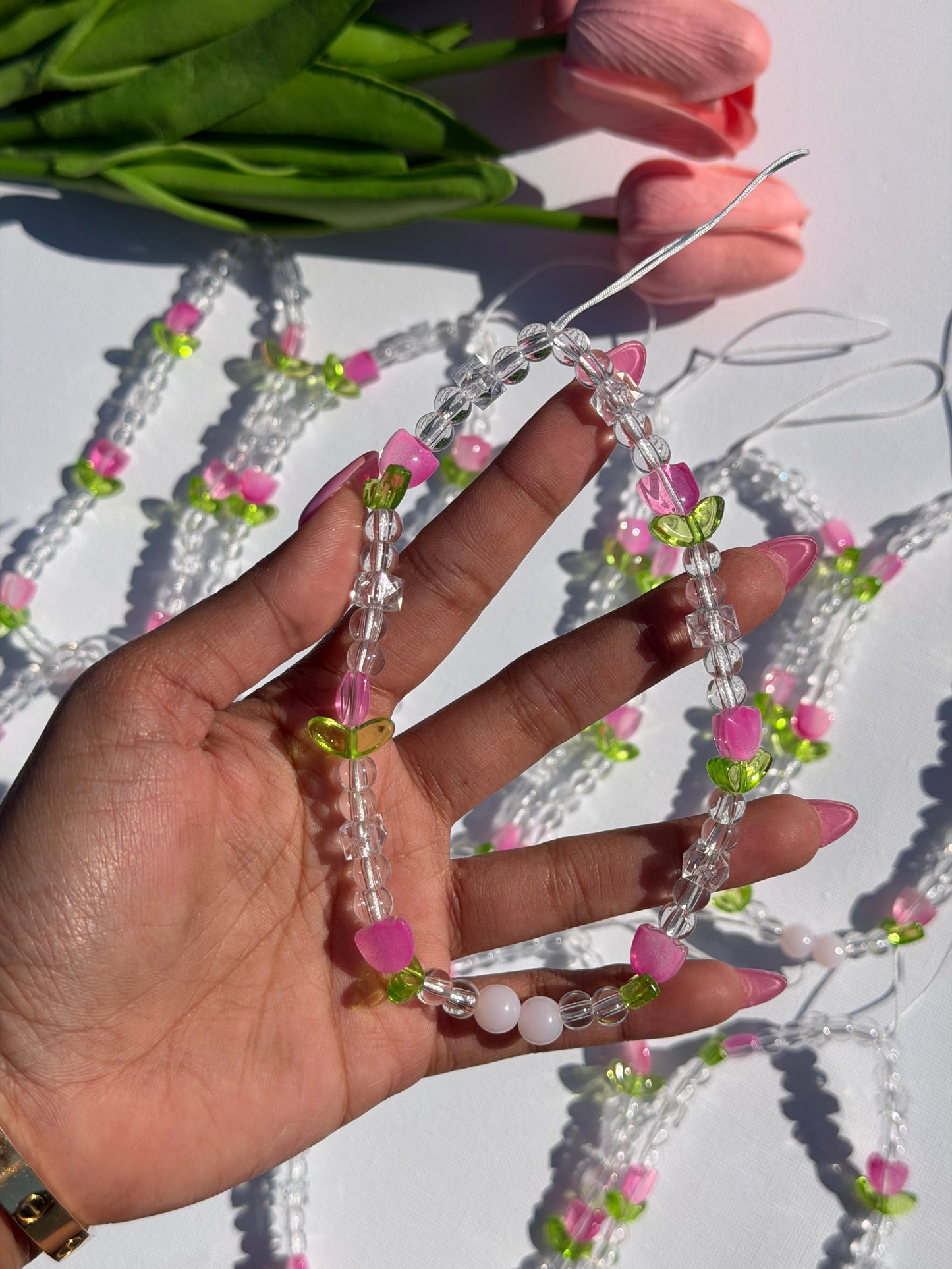 Beaded phone charm with clear, pink tulip, and green leaf beads, held by a hand, against a white background with blurred pink tulips.