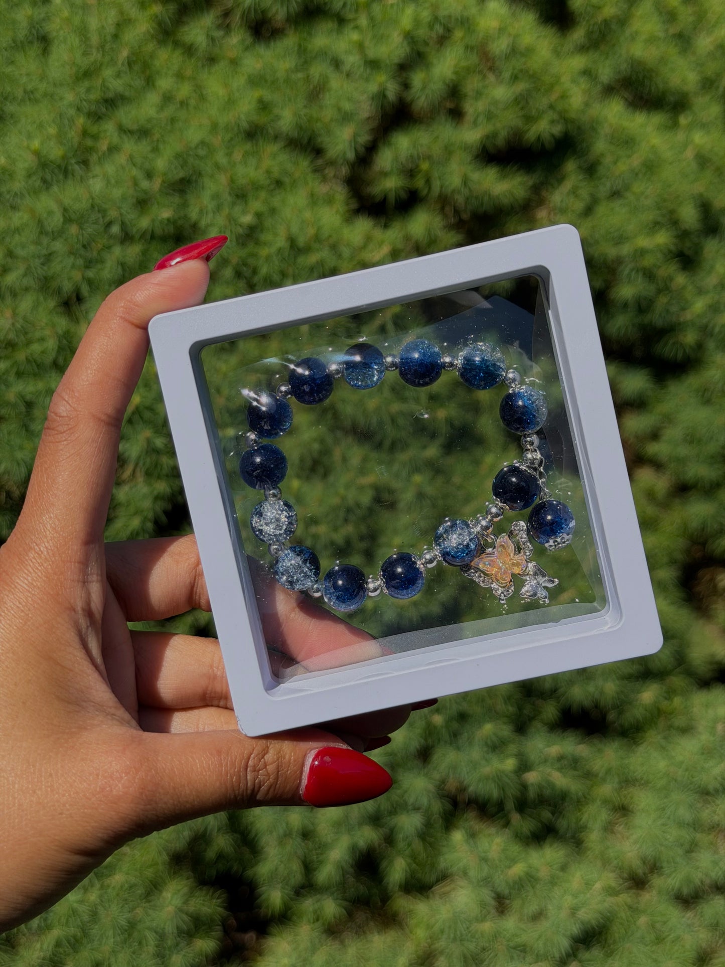 Blue beaded bracelet with silver accents and butterfly charm in clear display case, held in hand with red nails, green foliage background